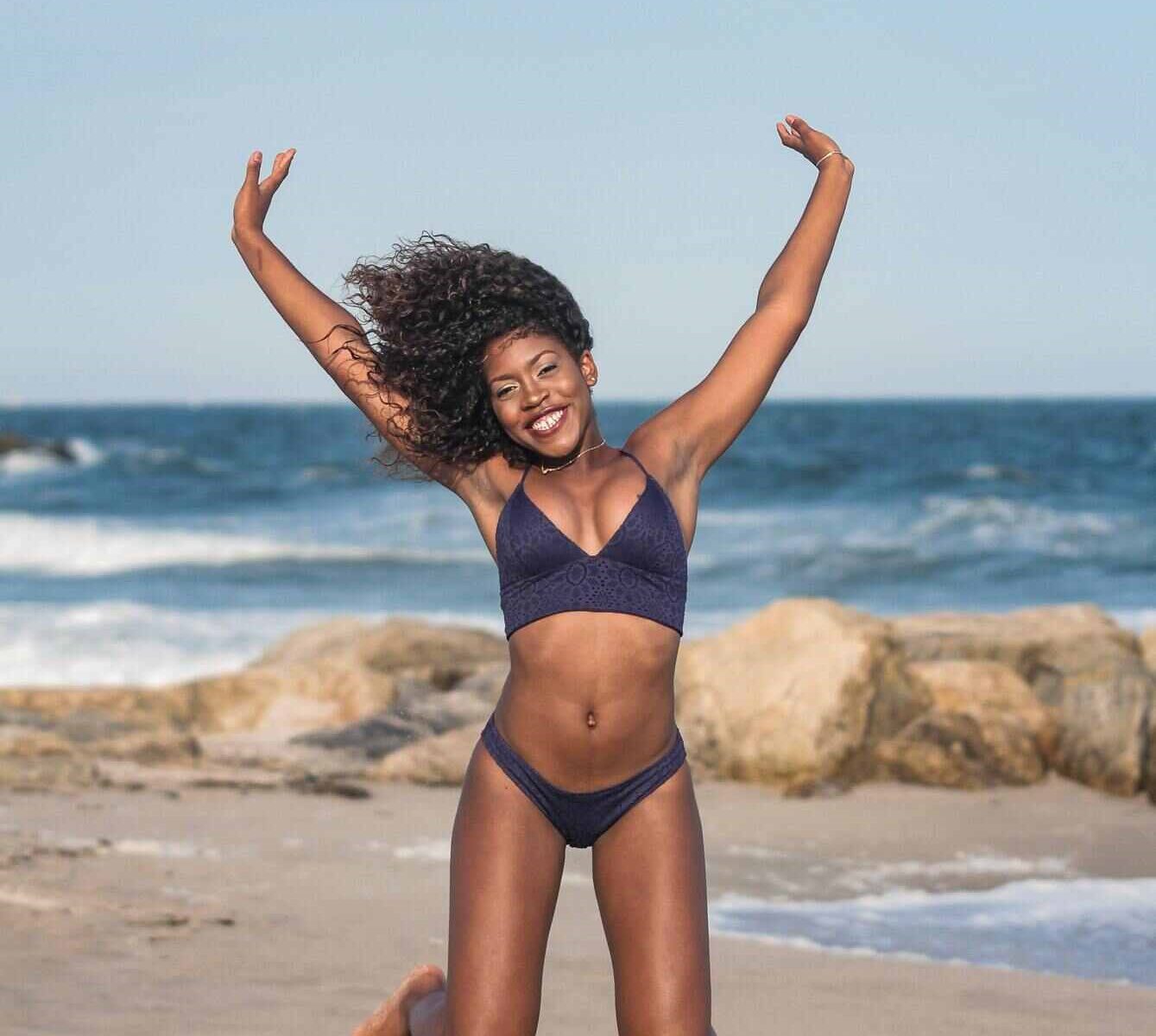 A woman in a bikini jumping. She's on a sandy beach and the blue sky is cloud free.