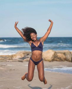 A woman in a bikini jumping. She's on a sandy beach and the blue sky is cloud free.