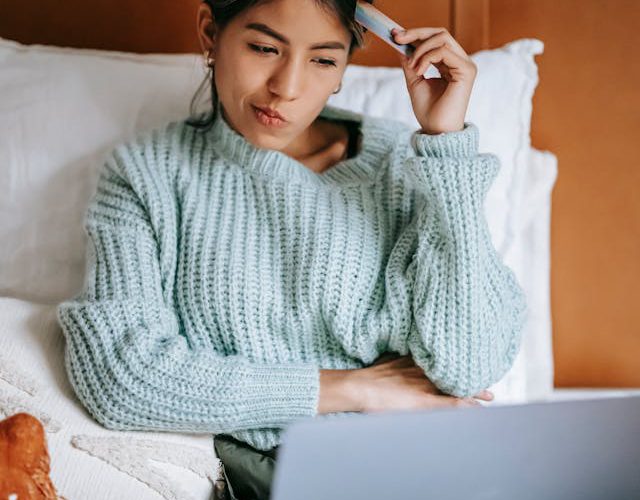 A woman sitting on her bed with her laptop on her lap. She is thinking while tapping her credit card against the side of her forehead.