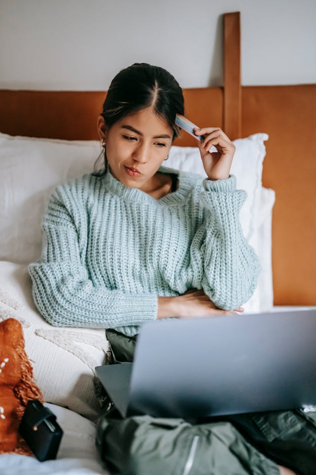 A woman sitting on her bed with her laptop on her lap. She is thinking while tapping her credit card against the side of her forehead.